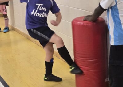 Child practices soccer dribbling around a red training dummy indoors.