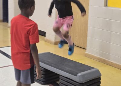 A child jumps over stacked gym steps while another watches.