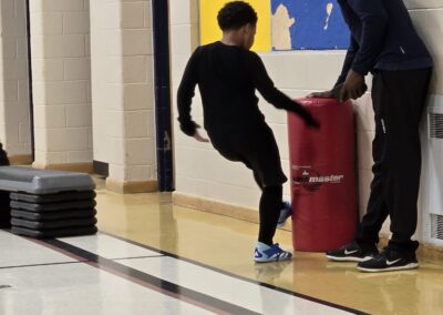 Child kicks a red trash bin while an adult stands nearby in a gym.