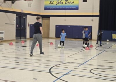 A coach instructs young kids playing soccer in a gymnasium.