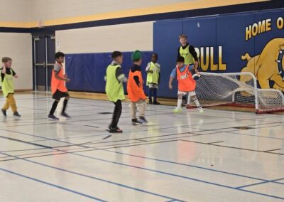 Children playing indoor soccer in a gymnasium wearing orange and green jerseys.