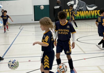 Two kids in soccer uniforms practicing indoors with a ball.