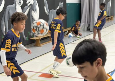 Children playing indoor soccer in a gymnasium wearing matching sports uniforms.