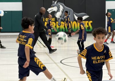 Kids playing soccer indoors with a coach guiding them.