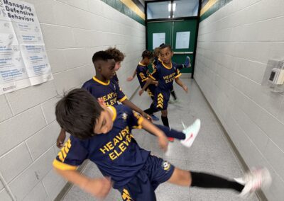 Kids in sports uniforms playing indoors, one kicking a ball.