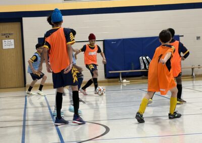 Kids playing indoor soccer in bright jerseys on a gym court.