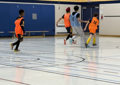 Youth indoor soccer game with players in orange and blue jerseys.