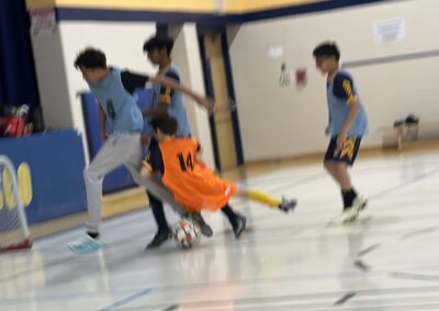 Children playing indoor soccer in a gym, one wearing an orange jersey.