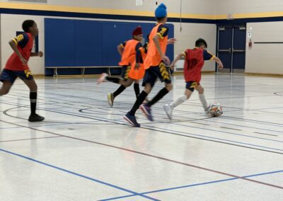 Youth indoor soccer game with players in orange and red jerseys.