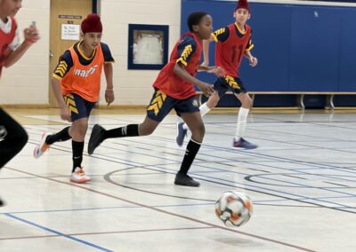 Three boys playing indoor soccer, wearing red and orange jerseys.