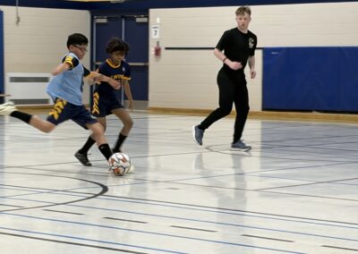 Two kids playing indoor soccer with a coach watching.