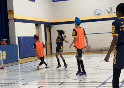 Kids playing basketball indoors wearing orange and blue jerseys.