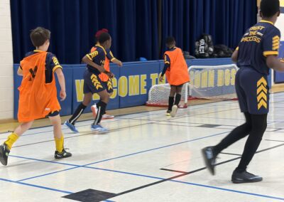 Children playing indoor soccer wearing orange bibs on a gym floor.