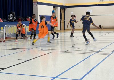Children playing indoor soccer wearing orange and blue jerseys.