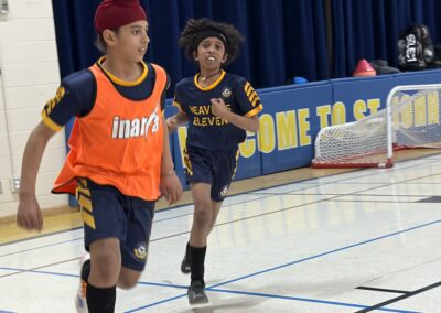 Two boys playing basketball indoors, one wearing an orange bib and the other in a navy jersey.
