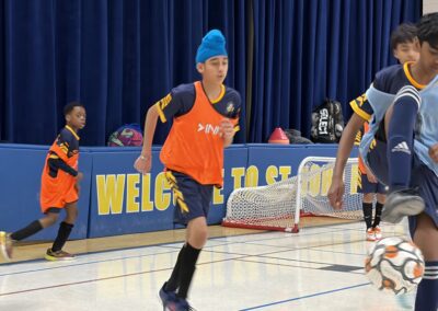Young boys playing indoor soccer in orange jerseys and blue hats.