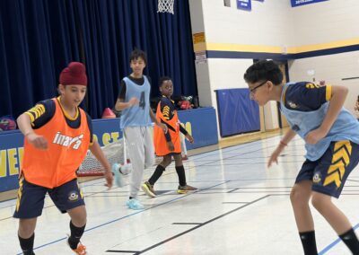 Children playing basketball indoors wearing orange and blue jerseys.