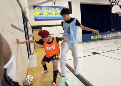 Two kids playing soccer indoors, one dribbling the ball.